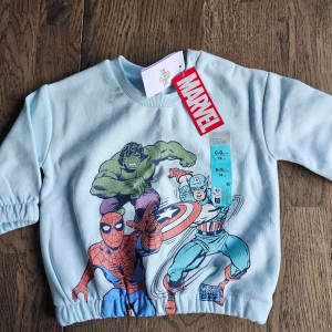 Ljusblå Marvel-tröja med superhjältar - Ljusblå långärmad tröja från Marvel x Primark med tryck av Hulken, Spiderman och Captain America på framsidan. Tröjan har resor i armar och nederkant och knappar på axeln. perfekt för små superhjältar.