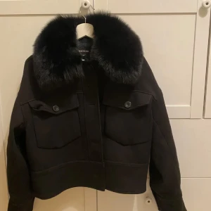 Xs Svart jacka med päls från Meotine - Snygg svart teddyjacka från Medotine med stor fluffig pälsimitation på kragen. Jackan har en croppad passform, två stora fickor med knappar framtill och knappar längs hela framsidan. Materialet är ullblandning och pälsdetaljen ger en lyxig vibe. Använd få gånger, Nypris 3000 :-