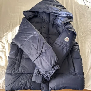 Mörkblå Moncler pufferjacka M - Säljer en mörkblå pufferjacka från Moncler med huva och klassisk logga på ärmen. Jackan är quiltad och har dragkedja samt knappar framtill. Perfekt för kalla dagar och riktigt snygg streetstil.