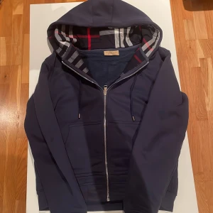 Marinblå Burberry hoodie med dragkedja - Säljer en marinblå hoodie från Burberry med klassiskt rutigt foder i huvan. Tröjan har hel dragkedja, broderad logga på ärmen och två fickor framtill. Perfekt för dig som vill ha en stilren och lyxig streetwear-look. Den är knappt använd så den är i nyskick/mycket bra skick. Kan gå ner i pris vid en snabb affär.