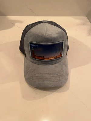 Grå truckerkeps med Stockholmstryck - Snygg grå mocka keps från Revenite ”säljs inte längre” med mesh på sidorna och ett stort tygmärke framtill som visar en kvällsbild av Stockholms stadshus och segelbåtar. Texten 'REVENITE POP UP '23' syns på märket. 