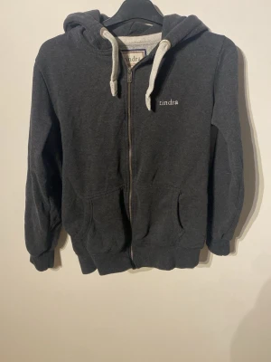 Mörkgrå hoodie med dragkedja Tindra - Mörkgrå hoodie från Tindra med vit dragsko i huvan och dragkedja framtill. Tröjan har två fickor fram och ribbade muddar vid ärmslut och nederkant. Perfekt för en avslappnad och stilren look.