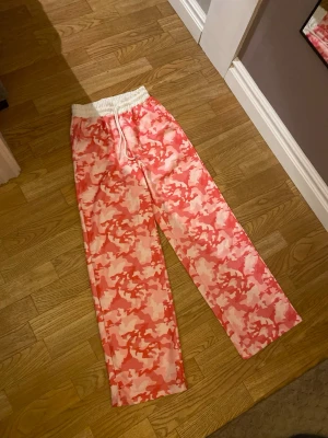 Rosa camo mjukisbyxor med snörning - Säljer ett par mjukisbyxor med rosa och vita camouflagemönster. Byxorna har bred resårmidja med snörning och raka ben för en chill och loose vibe. Perfekta för att hänga hemma eller dra till kompisar i.