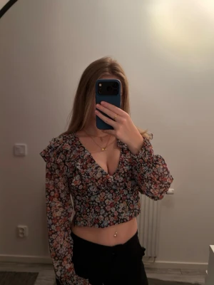 Blommig croppad blus med volang - Använd 1 gång. Passar även de med storlek s. Supersöt blommig croppad blus med volangdetaljer och v-ringning. Blusen har långa ärmar, öppen rygg med knytband och en dragkedja på sidan för att enklare kunna få av och på den.