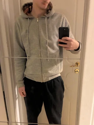 Burberry zip hoodie - Grå zip hoodie från Burberry med klassiskt rutigt mönster i huvan, fickor framtill och diskret logga på bröstet. Perfekt för dig som gillar stilrena och exklusiva plagg. Tröjan har en liten fläck på ena armbågen (se sista bilden). Hör gärna av dig vid frågor eller liknande🙏