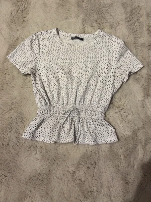 Vit prickig blus med knyt i midjan - Söt vit blus med svarta prickar och korta ärmar. Toppen har ett knytband i midjan och volangkant nedtill som ger en feminin siluett. Perfekt till jeans eller kjol för en trendig look.