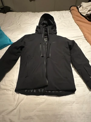 Svart skidjacka från Five Seasons - Svart skidjacka från Five Seasons med huva, flera dragkedjeförsedda fickor och snyggt quiltat foder. Jackan har lång ärm och diskret logga på bröstet. Perfekt för kalla dagar med sin isolerade insida och stilrena look. Storlek S passa även M också perfekt för att åka skidor eller snowboard 