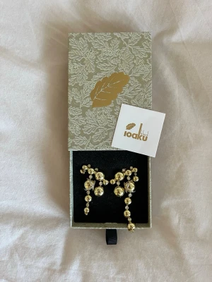 Guldiga statementörhängen från Ioaku - Unika örhängen från Ioaku i guldton med klotformade detaljer och gnistrande stenar💗 Kommer i originalask med snyggt mönster och logga. Aldrig använda! Org.pris: 999kr