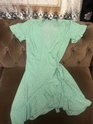 Grön omlottklänning med volang - Söt grön omlottklänning med små vita blommor, kort ärm och volangkant. Klänningen har knyt i midjan och en luftig, kort passform som ger en somrig känsla. Perfekt för dig som gillar en feminin och avslappnad stil.