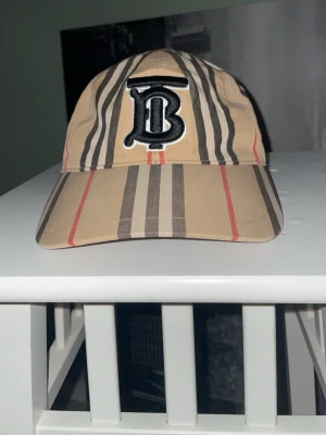 Beige keps med Burberry-mönster - Snygg beige keps från Burberry med klassiskt rutigt mönster i svart, vitt och rött. Stor broderad B-logga framtill och justerbar rem baktill med metallknappar. Perfekt accessoar för en trendig streetstyle-look. Serösa köpare bara