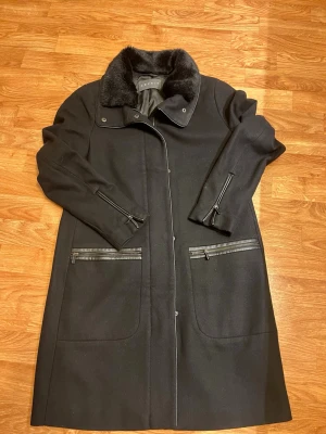 Svart kappa från Esprit med fuskpäls - Stilren svart kappa från Esprit med mjuk fuskpäls på kragen och snygga dragkedjedetaljer vid ärmar och fickor. Kappan har klassisk rak passform och är fodrad för extra komfort. Perfekt för kyliga dagar när du vill vara både varm och snygg.