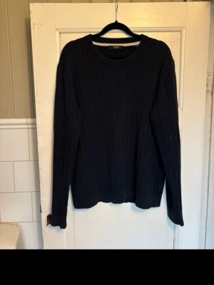 Mörkblå kabelstickad tröja Jack & Jones - Kabelstickad tröja från Jack & Jones i mörkblått. Klassisk rund halsringning och långa ärmar. Tröjan har ribbade muddar vid ärmslut och nederkant samt diskret logga vid höften. Perfekt för lager på lager och enkel att matcha. Liten defekt på ena armen, syns knappt vid användning