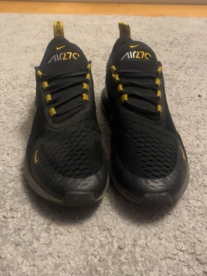 Nike Air270 svarta sneakers med gula detaljer - Säljer ett par nästan aldrig använda Nike Air270 sneakers i svart med gula detaljer och vita loggor. Skorna har ventilerande meshmaterial, snörning och en chunky sula med synlig Air-enhet i hälen. Perfekta för dig som vill ha en sportig och modern look.