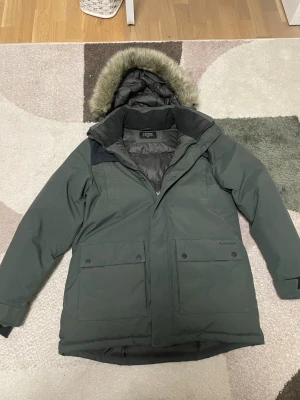 Grön parkasjacka från Everest” pris kan diskuteras” - Mörkgrön parkasjacka från Everest med avtagbar huva och fuskpälsdetalj. Jackan har stora framfickor med knappar och dragkedja framtill. Perfekt för kalla dagar med sin vadderade insida och klassiska look.