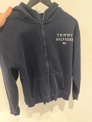 Marinblå hoodie Tommy Hilfiger - Snygg marinblå hoodie från Tommy Hilfiger med dragkedja framtill och fickor. Märkets logga broderad på bröstet och en vit/röd rand på huvan. Tillverkad i mjukt bomullsmaterial och har ribbade muddar vid ärmslut och nederkant.