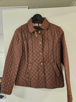 Brun quiltad vindjacka med krage - Snygg brun vindjacka med quiltat mönster och klassisk krage. Jackan har guldfärgade knappar framtill och två fickor med dragkedja. Perfekt för dig som vill ha en stilren och tidlös look.