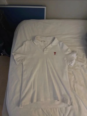 Vit pikétröja från Ami Paris - Klassisk vit pikétröja från Ami Paris med röd broderad logga i form av ett hjärta och bokstaven A på bröstet. Tröjan har krage, korta ärmar och knappar vid halsen. Perfekt till jeans eller shorts för en clean look.