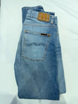 Blå jeans från Nudie Jeans w 31 L 30 - Säljer ett par klassiska blå jeans från Nudie Jeans med ljus tvätt och kontrastsömmar. Jeansen har en bakficka med Nudie-logga och patch i läder bak i midjan. Perfekta för en avslappnad och trendig look.W 31 L 30