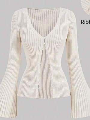 Vit ribbad kofta med v-ringning - Trendig vit ribbad kofta med djup v-ringning och utsvängda ärmar. Koftan har smala knappar framtill och är figurnära i modellen. Perfekt för dig som gillar en stilren och modern look.