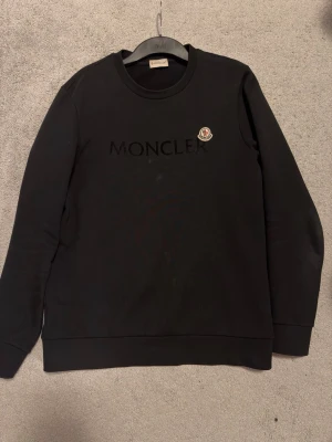 Svart Moncler sweatshirt  - Snygg svart sweatshirt från Moncler med broderad logotyp på bröstet och diskret Moncler-text framtill. Tröjan har rund halsringning och långa ärmar. Perfekt för en stilren och trendig look.