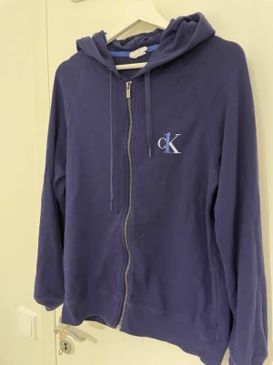 Mörkblå hoodie från Calvin Klein - Mörkblå hoodie från Calvin Klein med dragkedja framtill och klassisk huva. På bröstet finns en vit och blå CK-logga. .