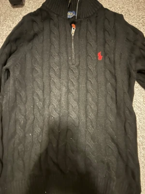 Svart kabelstickad tröja Polo Ralph Lauren - Svart kabelstickad tröja från Polo Ralph Lauren med halv dragkedja och röd broderad logga på bröstet. Tröjan har hög krage och är tillverkad i mjukt material, perfekt för kyliga dagar.