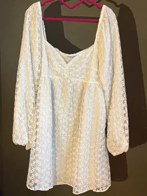 Vit blommig kortklänning med spets NAKD - Söt vit kortklänning med blommigt spetsmönster och långärmade puffärmar. Klänningen har en v-ringad urringning och är tillverkad i ett lätt, luftigt material som passar perfekt till varma dagar. Romantisk och trendig look med fina detaljer, perfekt för studenten. Köpt från NAKD och har endast använts en gång🐚🤍