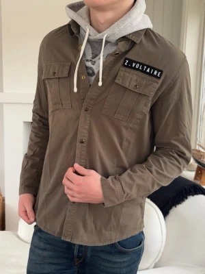 Olivgrön overshirt från Zadig & Voltaire - Snygg olivgrön overshirt från Zadig & Voltaire med två bröstfickor och knappstängning framtill. Patch med logga på vänster bröst. Skön passform och klassisk krage, perfekt att bära över hoodie eller t-shirt.
