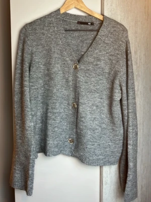 Grå kofta från 157 med knappar - Mysig grå kofta från 157 med tre stora knappar framtill. Koftan har v-ringning, lång ärm och är stickad i ett mjukt material. Perfekt att slänga över en t-shirt eller topp för en chill look.