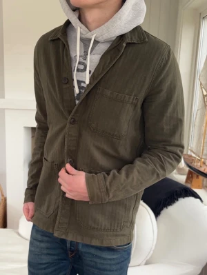 Olivgrön overshirt med knappar - Stilren olivgrön overshirt med klassisk krage och tre framfickor. Overshirten har knäppning framtill och är tillverkad i ett mjukt bomullstyg. Perfekt att bära över en hoodie för en avslappnad look.