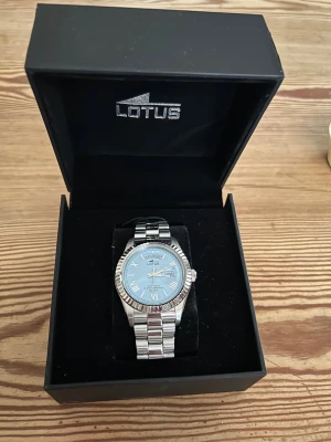 Klocka lotus silverfärgad  - Stilren klocka från Lotus med silverfärgat armband i metall och ljusblå urtavla. Klockan har romerska siffror, dag- och datumvisning samt räfflad boett. Perfekt accessoar för dig som gillar klassisk men modern stil. Ny skick har endast används ett få tal gånger. Köpt på ur och Penn ( kvitto finns! )                                 kan gå ner i pris vid snabb affär!!!              Original ask och extra länkar ingår