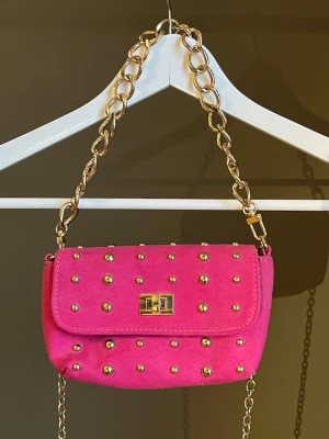 Rosa axelväska med guldkedja - Snygg rosa axelväska med guldfärgade nitar och kedja. Väskan har ett lock med vridlås i guld och är tillverkad i ett mjukt material som liknar mocka. Perfekt för dig som vill sticka ut med en färgglad accessoar. Köpt från Nelly, aldrig använt🎀