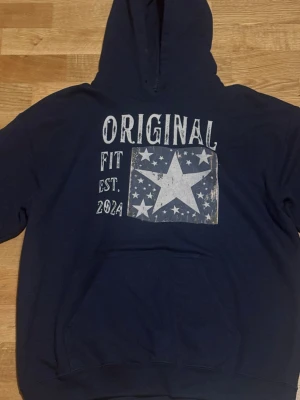 Mörkblå SCANDI hoodie  - Mörkblå hoodie från SCANDI med stort vitt tryck framtill med texten 'ORIGINAL FIT EST. 2024' och en stor stjärna omgiven av mindre stjärnor. Klassisk hoodie och huva. Perfekt för en avslappnad och cool stil. Storlek M Inga defekter dock inga snören använt fåtal gånger 