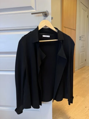 Svart kavaj från Mango - Stilren svart kavaj från Mango med klassisk krage och långa ärmar. Kavajen har axelklaffar och knappar vid ärmsluten för en extra detalj. Perfekt för dig som vill ha en enkel men snygg look.