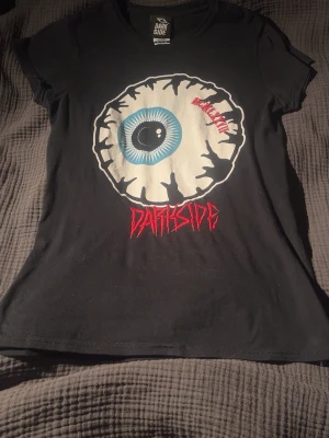 Svart t-shirt med ögonmotiv från Darkside - Cool svart t-shirt från Darkside med ett stort grafiskt ögonmotiv i vitt, blått och svart på bröstet. Under motivet står det 'Darkside' i röd text. T-shirten har korta ärmar och en klassisk passform. Perfekt för dig som gillar alternativ stil. Storleken säger M men skulle säga att den sitter mer som S