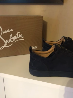 Blåa sneakers från Christian Louboutin - Stilrena svarta sneakers från Christian Louboutin med klassisk röd sula och ovandel i mocka. Skorna har rund tå, snörning och diskret logga på sidan. Perfekta för dig som gillar exklusiva detaljer och tidlös design.
