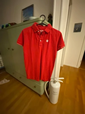 Röd piké från 8848 Altitude - Snygg röd pikétröja från 8848 Altitude med klassisk krage och knappar framtill. Tröjan har korta ärmar och en vit siffra 8 på bröstet. Perfekt för en sportig och avslappnad stil.