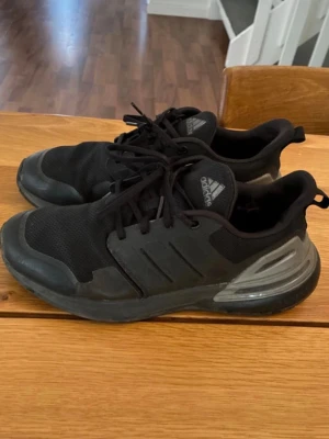 Svarta Adidas sneakers med snörning - Säljer ett par svarta Adidas sneakers med klassisk mesh- och syntetöverdel, samt tydliga tre ränder på sidan. Skorna har snörning och en bekväm, vadderad häl. Perfekta för dig som gillar sportig stil och vill ha något som funkar till träning eller chill.