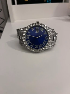 Silverfärgad klocka med blå urtavla - Säljer en riktigt blingig klocka med silverfärgat armband och blå urtavla. Klockan är täckt med gnistrande stenar runt boetten och på länken. Romerska siffror och datumvisning på urtavlan. Perfekt för dig som gillar att sticka ut och vill ha något extra på handleden.