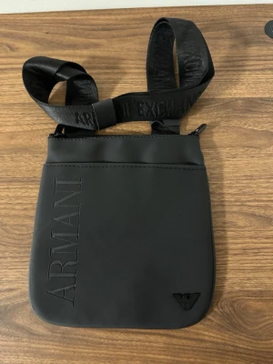 Svart axelväska från Armani Exchange - Snygg svart axelväska från Armani Exchange med justerbart axelband där loggan är invävd. Väskan har stilren design med präglad ARMANI-text på framsidan och diskret logotyp i hörnet. Perfekt för att bära det viktigaste med stil.
