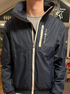 Marinblå vindjacka från Sail Racing - Snygg marinblå vindjacka från Sail Racing med vita detaljer och dragkedjor. Jackan har hög krage, ribbade muddar och en bröstficka med dragkedja. Tillverkad i lätt polyester som passar perfekt för blåsiga dagar.