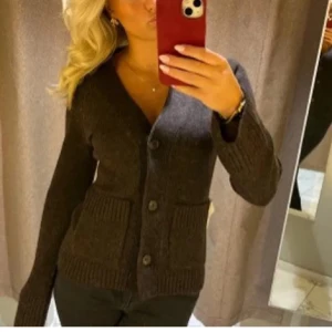 Mörkbrun kofta från Wera - Mysig mörkbrun kofta från Wera med v-ringning, tre knappar framtill och två stora fickor. Koftan har långa ärmar och ribbade muddar vid ärmslut och nederkant. Perfekt för lager på lager och enkel att matcha med olika outfits. 