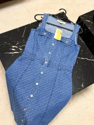 Blå jeansklänning Michael Kors - Snygg blå jeansklänning från Michael Kors med knappar framtill och diskret mönster. Klänningen har breda axelband, fickdetaljer på bröstet och markerad midja. Perfekt för dig som gillar en trendig och avslappnad look.