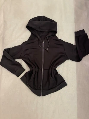 Mörk grå zip hoodie med huva - En stilren mörk grå hoodie med dragkedja framtill och huva. Hoodien har två fickor på sidorna och ribbade muddar vid ärmsluten. Perfekt för en avslappnad look och enkel att matcha med allt. Materialet känns mjukt och bekvämt. Den är i storlek L och passar perfekt för dig som gillar oversized hoodies. 