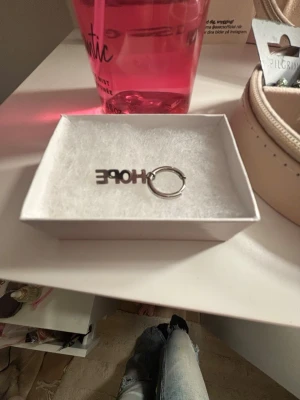 Silverfärgat örhänge med HOPE-text ifrån Edblad  - Coolt örhänge i silverfärg med texten 'HOPE' som hänge. Ringen är rund och enkel, medan texten ger en personlig touch. Perfekt accessoar för dig som gillar statement-smycken och vill sticka ut lite extra. Obs endast 1 öronhänge!