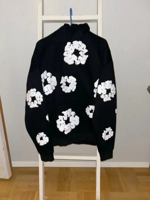 Svart hoodie med vita blommor - Cool svart hoodie med stora vita blommönster över hela tröjan. Oversized passform med huva och känguruficka framtill. Perfekt för dig som gillar streetwear och vill sticka ut med en unik design. Mjuk bomullskänsla och ribbade muddar vid ärmslut och nederkant.