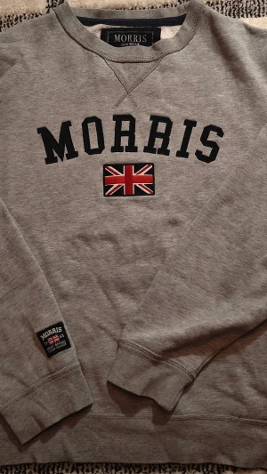 Grå sweatshirt från Morris - Snygg grå sweatshirt från Morris med svart text och en broderad brittisk flagga på bröstet. Tröjan har rund halsringning och ribbade muddar vid ärmslut och nederkant. Perfekt för en avslappnad och stilren look.