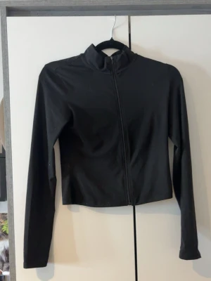 Svart ziptröja från SHEIN - Svart långärmad ziptröja från SHEIN i stretchigt material med 95% polyester och 5% elastan. Tröjan har hög krage och en smal, croppad passform. Perfekt för en stilren och enkel look.