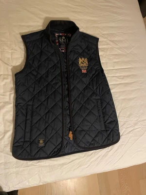 Mörkblå quiltad dunväst från Morris - Stilren mörkblå dunväst från Morris med quiltat mönster, ståkrage och broderad logga på bröstet. Västen har två fickor framtill och snygga detaljer med brittisk flagga på insidan. Perfekt för lager-på-lager och en klassisk look. Använt nån gång
