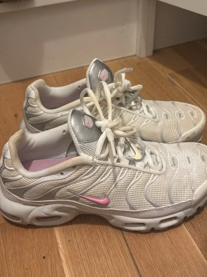 Nike Air Max Plus vita och rosa sneakers - Nike Air Max Plus sneakers i vitt med silverdetaljer och rosa inslag. Skorna har mesh- och syntetmaterial, klassisk snörning och synlig Tn-logga under sulan. Snyggt vågmönster på ovandelen och rosa swoosh på sidan. Perfekta för dig som gillar sportig stil. Dom är igentligen helt nya bara använda en gång men råka tappa kaffe på dom!